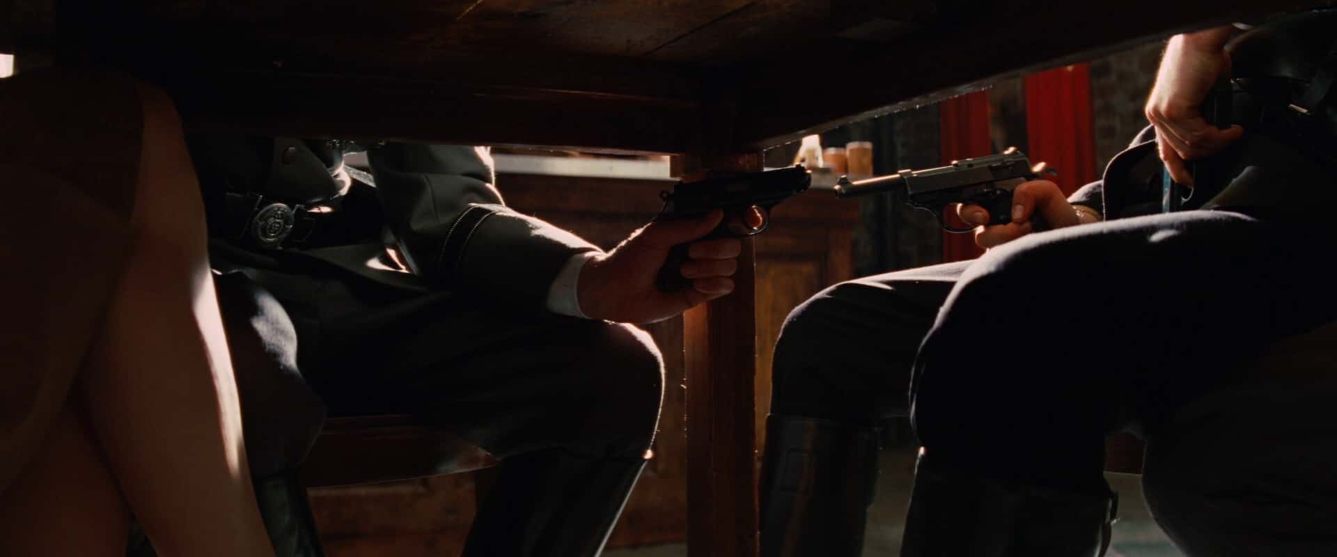 abaixo de uma mesa, dois homens seguram armas um contra o outro