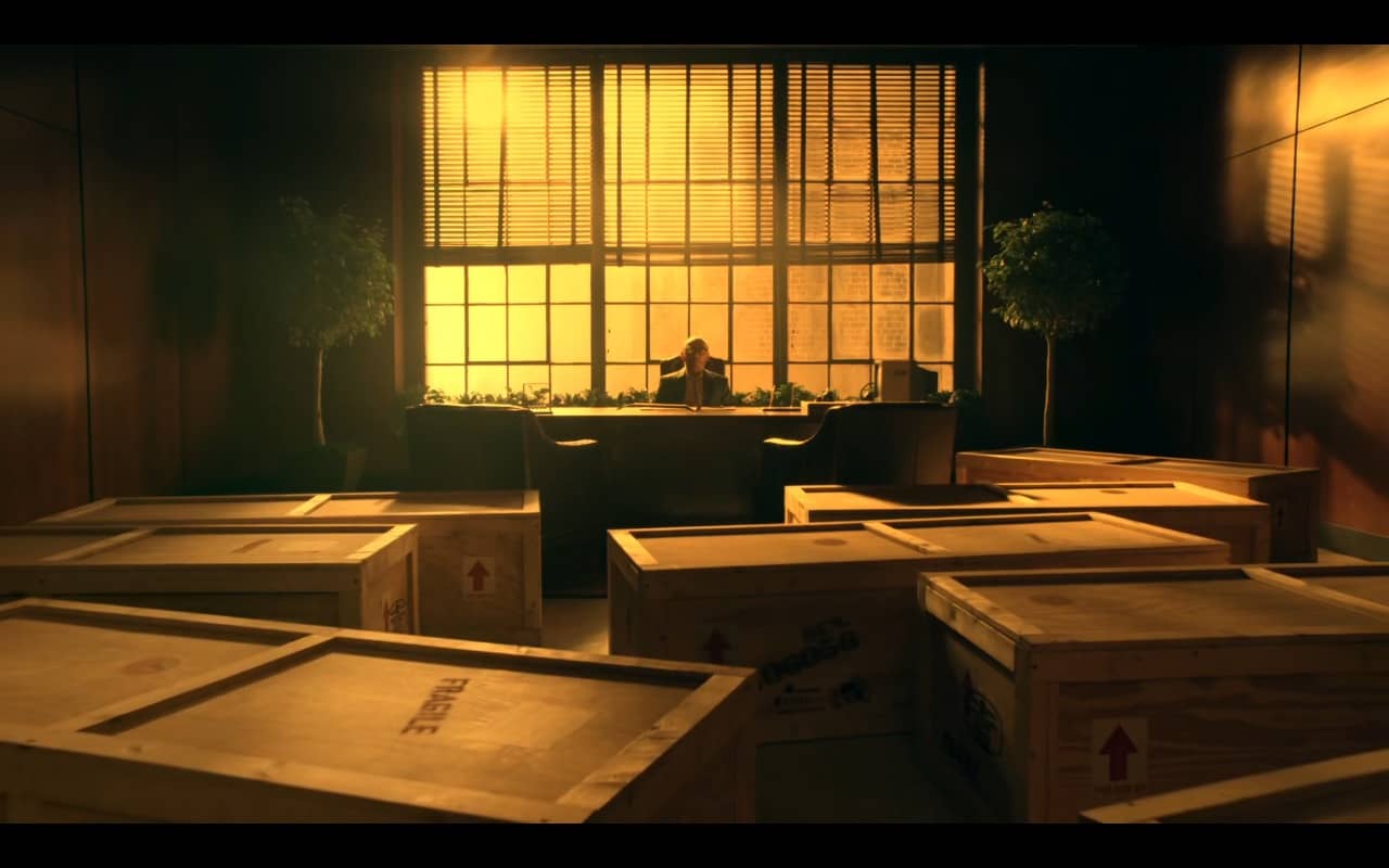 numa grande sala, diversos caixotes, parecendo caixões funerários, dispostos em frente a mesa de um homem de meia idade