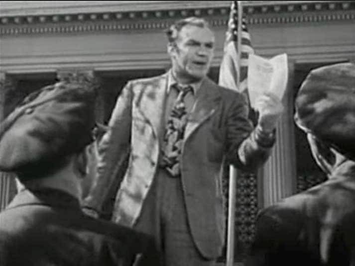 homem falando com fervor , em frente a uma bandeira americana e agitando um papel, enquanto observado por um público em uma praça