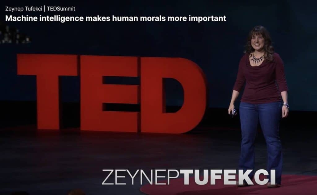 Uma mulher em um palco, vista de corpo inteiro, vestida com blusa roxa e calça jeans, com os braços ao seu lado, estendidos. Ao fundo do palco, as letras TED esculpiadas com cerca de um metro de altura cada, na cor vermelha.