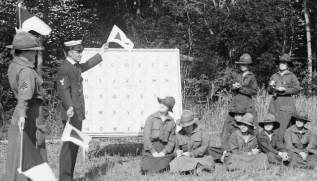 5 uniformed women seated and two on foot to the right, they are looking to the left, where a man and another womenare showingyon some flags. Between them a white board with some letters. Behind them, some trees.