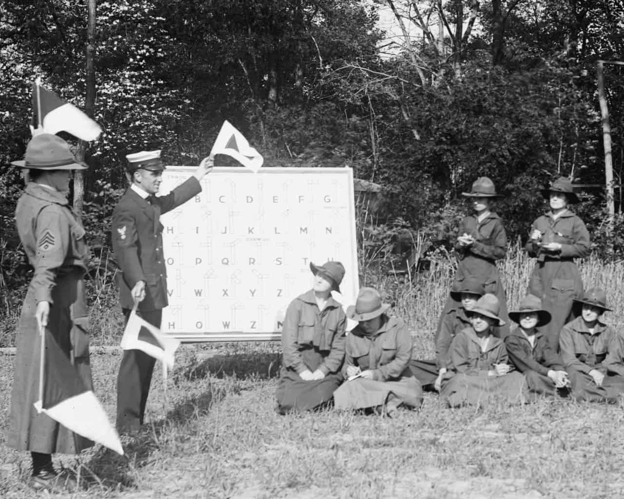 5 uniformed women seated and two on foot to the right, they are looking to the left, where a man and another womenare showingyon some flags. Between them a white board with some letters. Behind them, some trees.